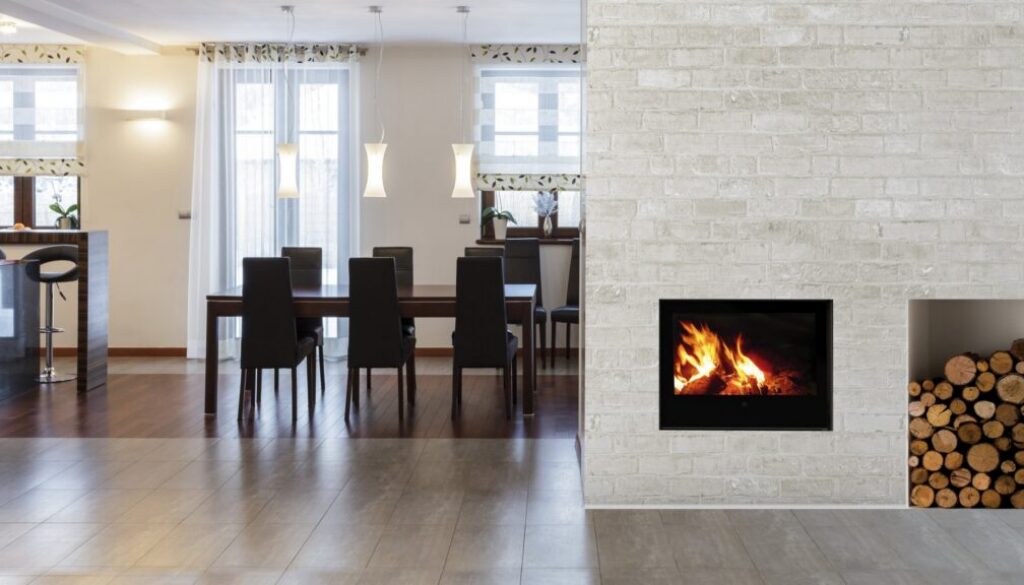 Holzofen mit Zentralheizung: wasserführender Holzofen mit Feuer in einer hellen Wandverkleidung, daneben eine Holzlege im offenen Wohnraum
